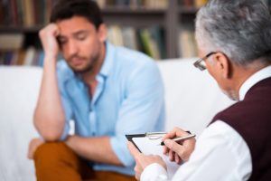Sad young man talking with psychologist. Psychologist taking notes. There are many books in psychologist office